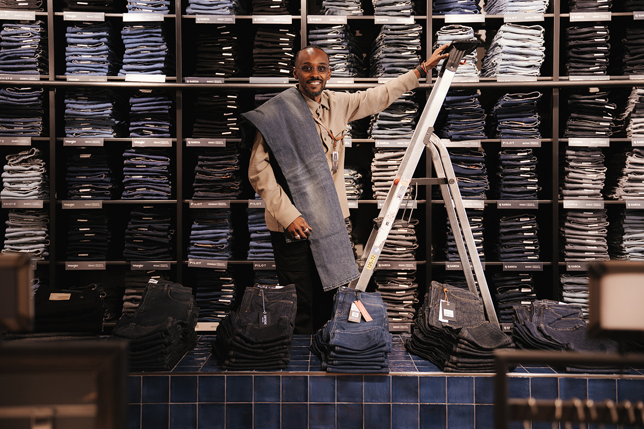 Man staat voor een wand met opgestapelde spijkerbroeken en een ladder.